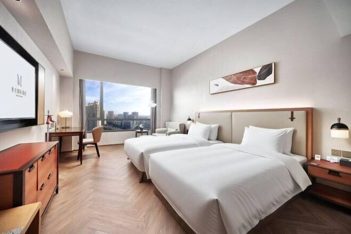 Ein Hotelzimmer mit zwei Betten, einem Schreibtisch, einem Sessel, einem Fernseher und Blick auf die Stadt.