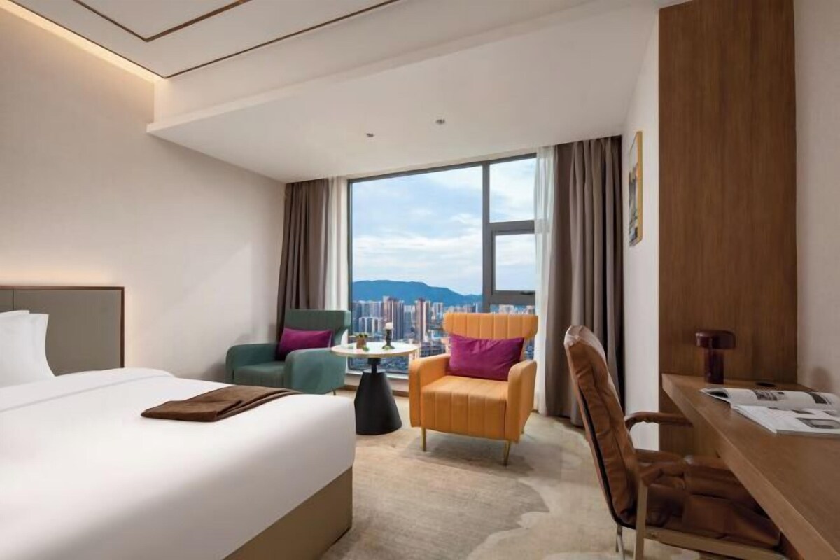 A hotel room with a large bed, a desk, two chairs, a view of the city, and a large window.