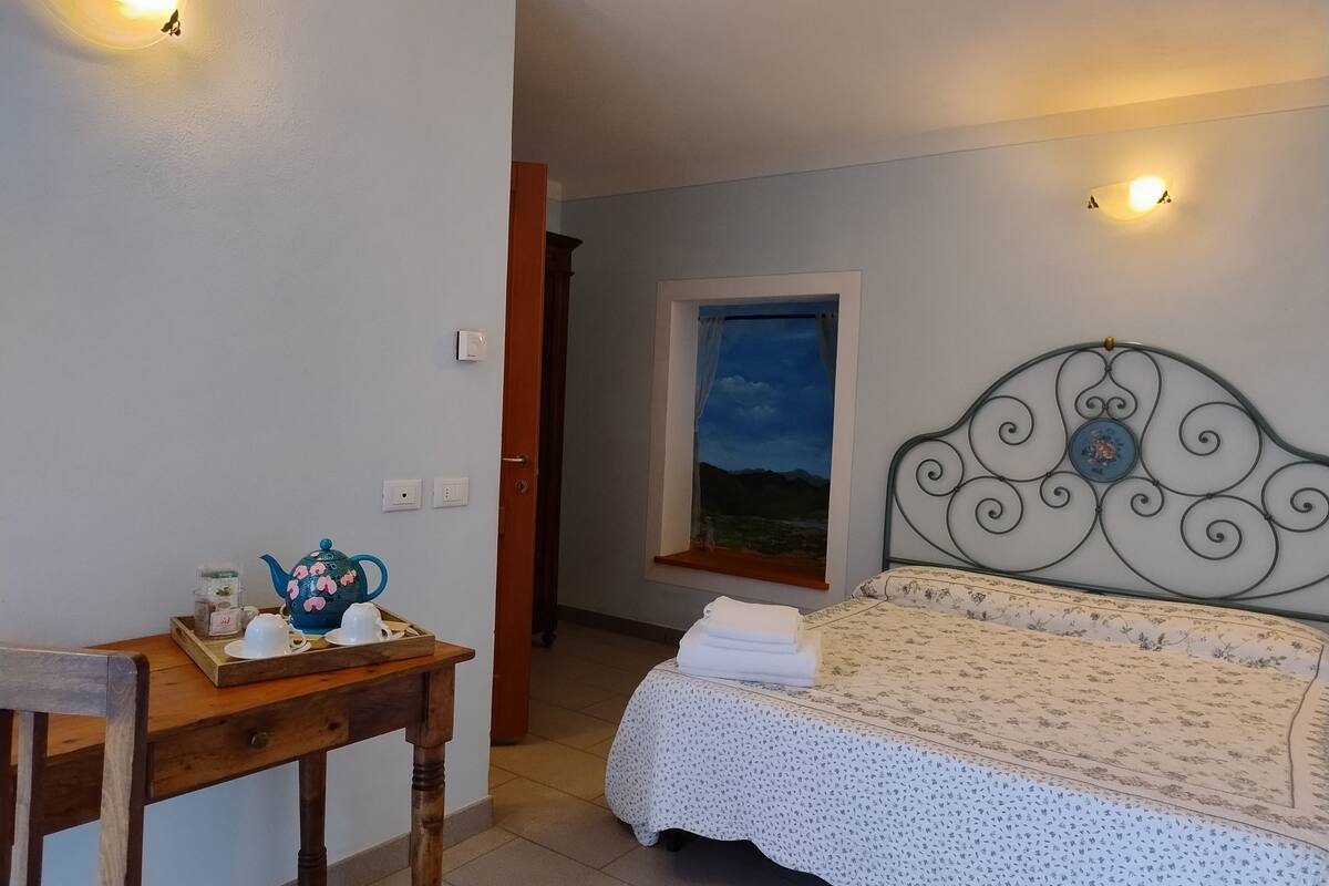A bedroom with a bed, a wooden table with a teapot, and a window with a view of the sky.