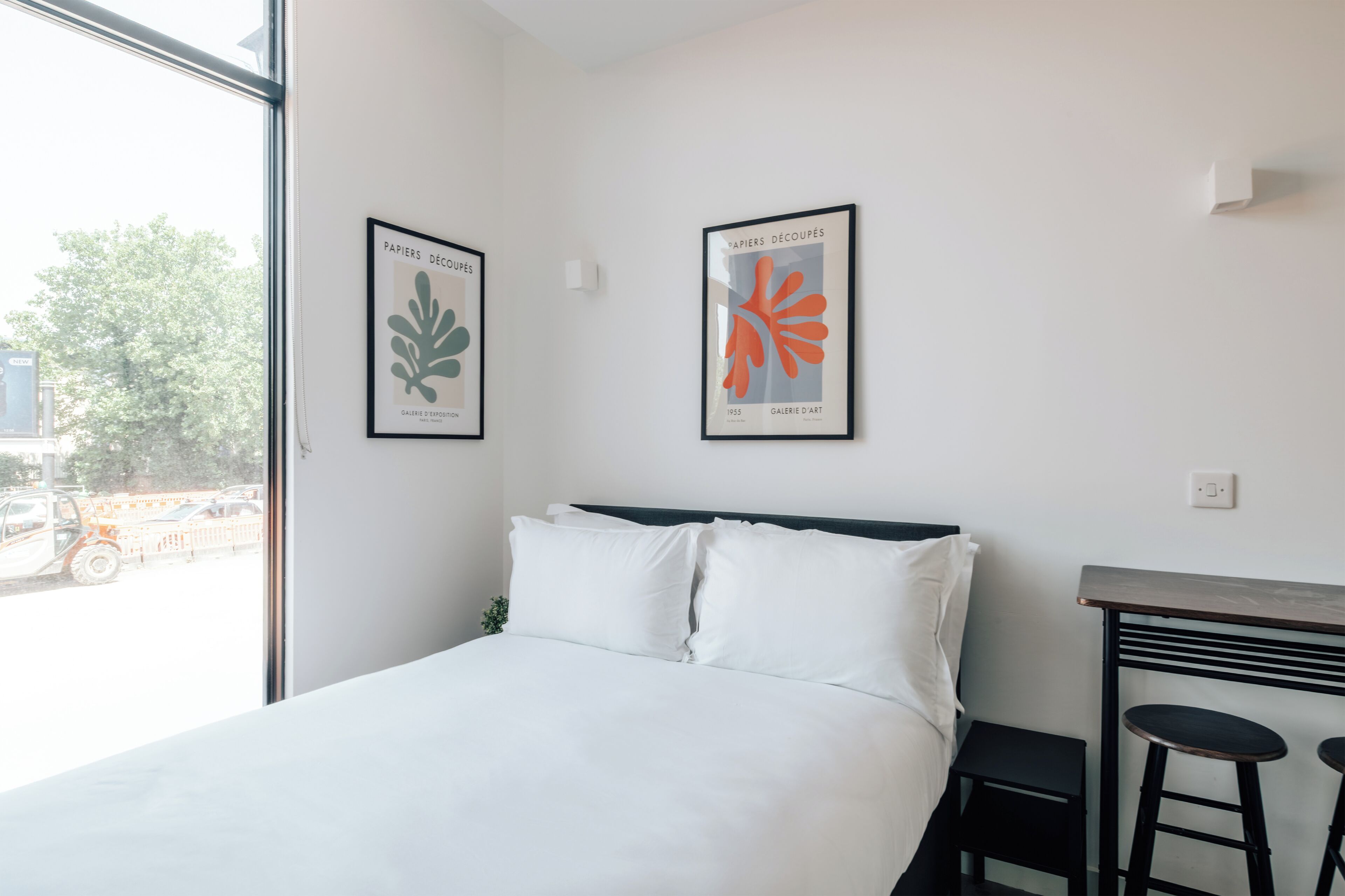 Een hotelkamer met een bed, twee ingelijste kunstwerken, een raam en een klein tafeltje met twee krukken.