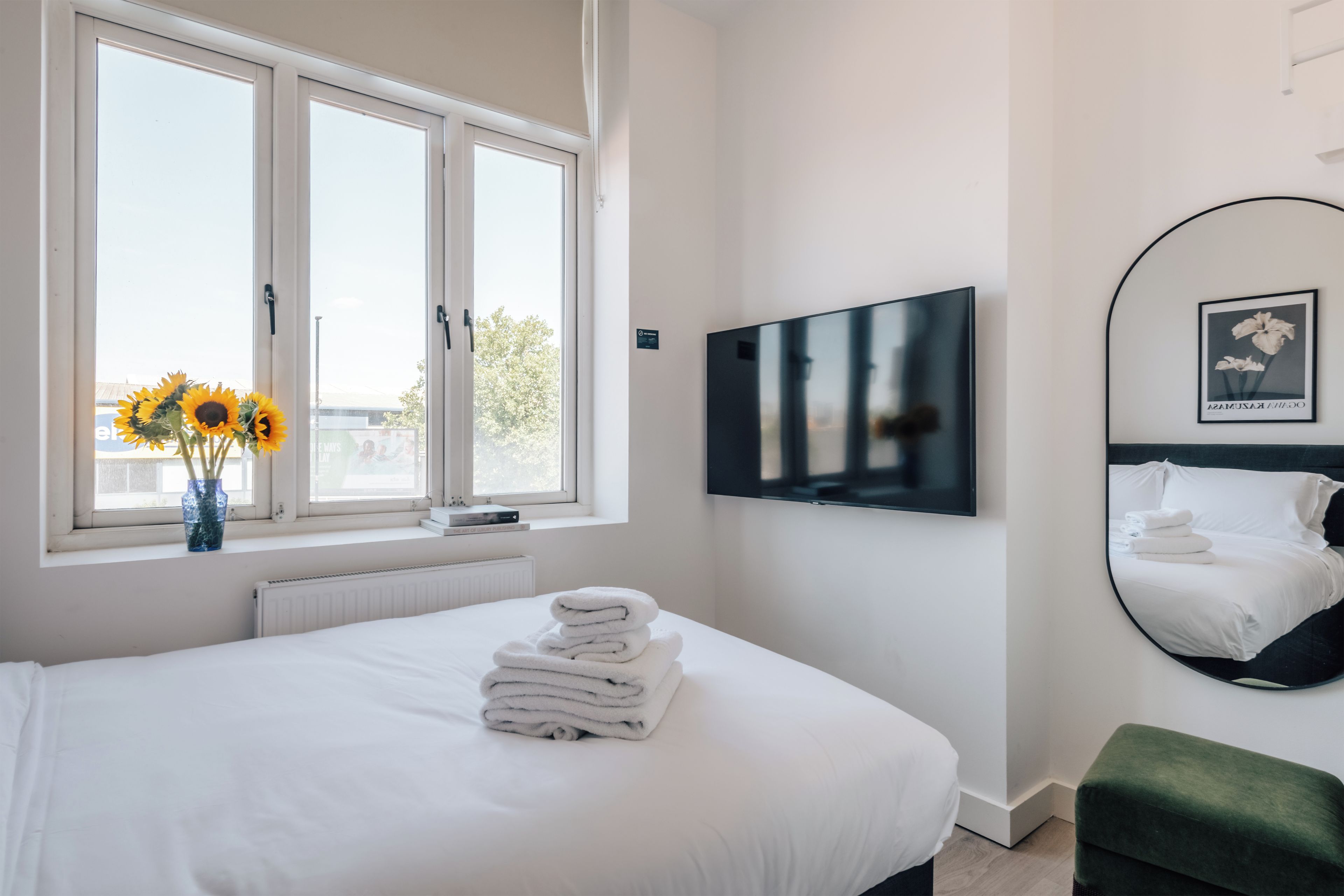 A hotel room with a bed, a TV, a mirror, and a window with sunflowers.