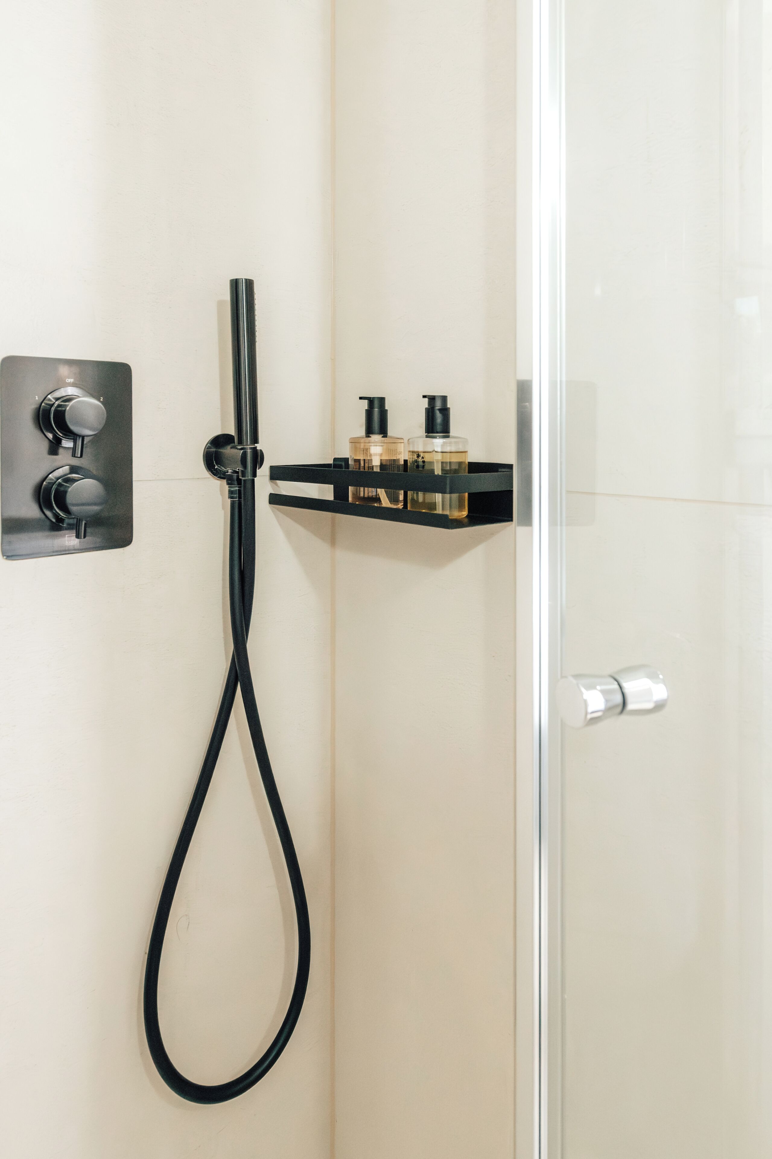 A shower with a handheld hose, a control panel, and a shelf with toiletries.