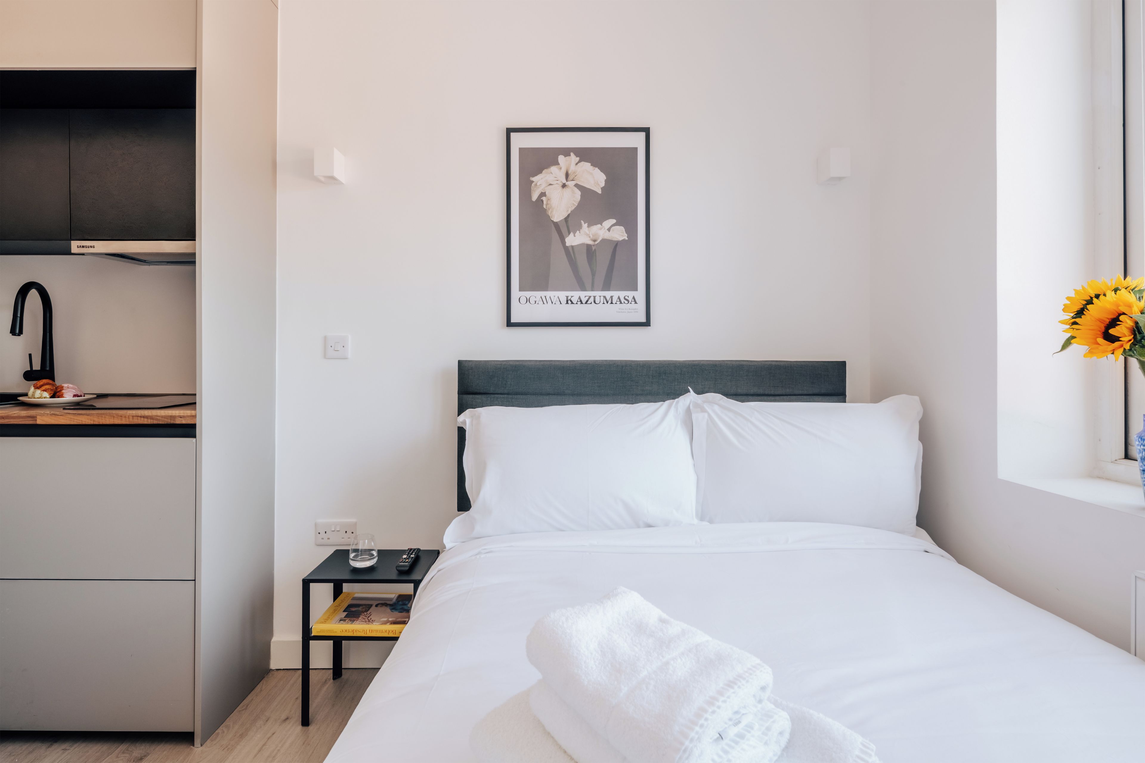 A hotel room with a bed, a nightstand, a framed picture on the wall, a window with a sunflower, and a kitchenette area.