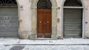 A building facade with a central wooden door, two arched windows, and a closed metal shutter to the right.