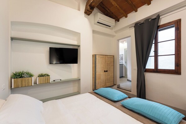 A bedroom with a bed, a TV mounted on the wall, a wooden wardrobe, and a window with curtains.