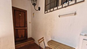 Ein kleiner Raum mit Holztür, einer Bank, einem Tisch, einem an der Wand befestigten Regal und einem Fenster mit Gitterstäben.