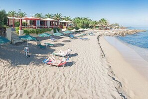 A beach with sun loungers and umbrellas, a building with a red roof, and palm trees. - Summer Home of Skaleta beach (9JP7+RM9 Skaleta)