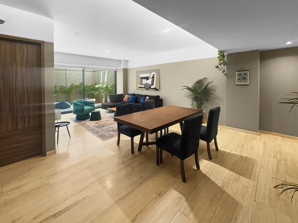 Un salon moderne avec une table à manger en bois, des chaises noires et un canapé agrémenté de coussins bleus. Une grande fenêtre offre une vue sur la verdure environnante.