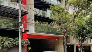 Un edificio de varios pisos con fachada roja y gris, una zona de estacionamiento techada y un coche estacionado al frente.
