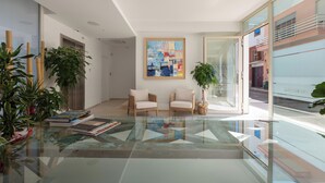 Una sala de estar moderna con una mesa de centro de vidrio, dos sillones, un cuadro abstracto de gran tamaño y plantas en macetas.