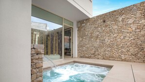 Un área de spa con jacuzzi, paredes de piedra y puerta de vidrio que da a una piscina.