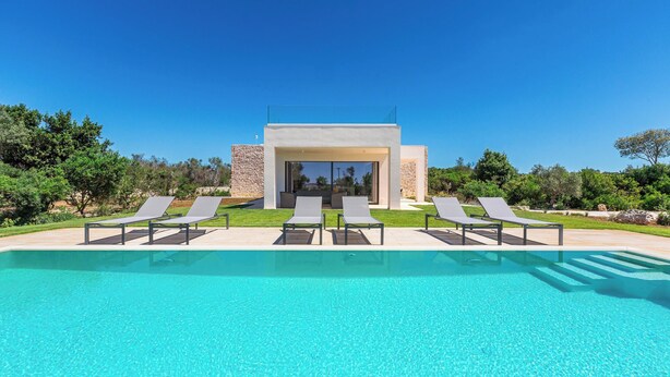 Un espace piscine moderne avec une eau d’un bleu limpide, entouré de chaises longues et d’un bâtiment en pierre doté de portes vitrées.