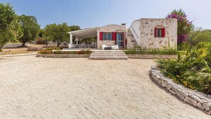 A stone building with a covered patio and red shutters, surrounded by gravel and plants.
