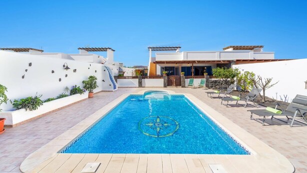 A swimming pool with a geometric design, surrounded by lounge chairs and potted plants, with a modern building in the background. - Villa Arisen by Villa Plus (Playa Blanca)