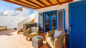 A covered outdoor area with two beige sofas, a small table with a plant, and a stone wall. There is a blue door and a staircase leading to an upper level. - Villa Arisen by Villa Plus (Playa Blanca)