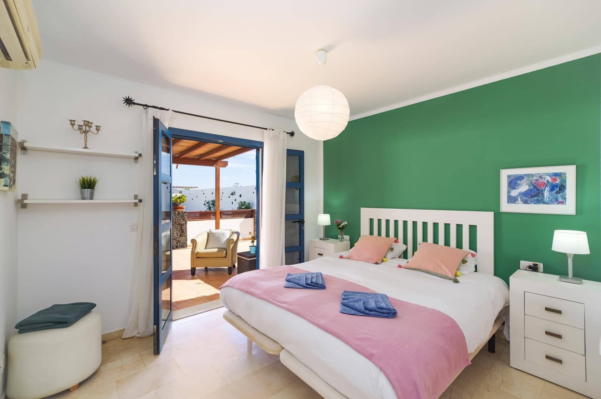 A bedroom with a bed, bedside tables, a nightstand, a chair, a potted plant, and a view through an open door to an outdoor area with a chair and a table.