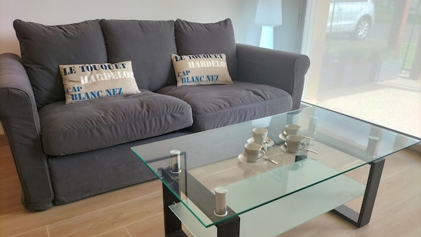 A modern living room with a grey sofa, two decorative pillows, and a glass coffee table with four white cups and saucers.