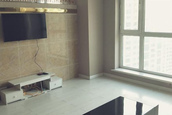 A modern living room with a flat-screen TV mounted on the wall, a white entertainment unit, and a large window providing a view of buildings outside.