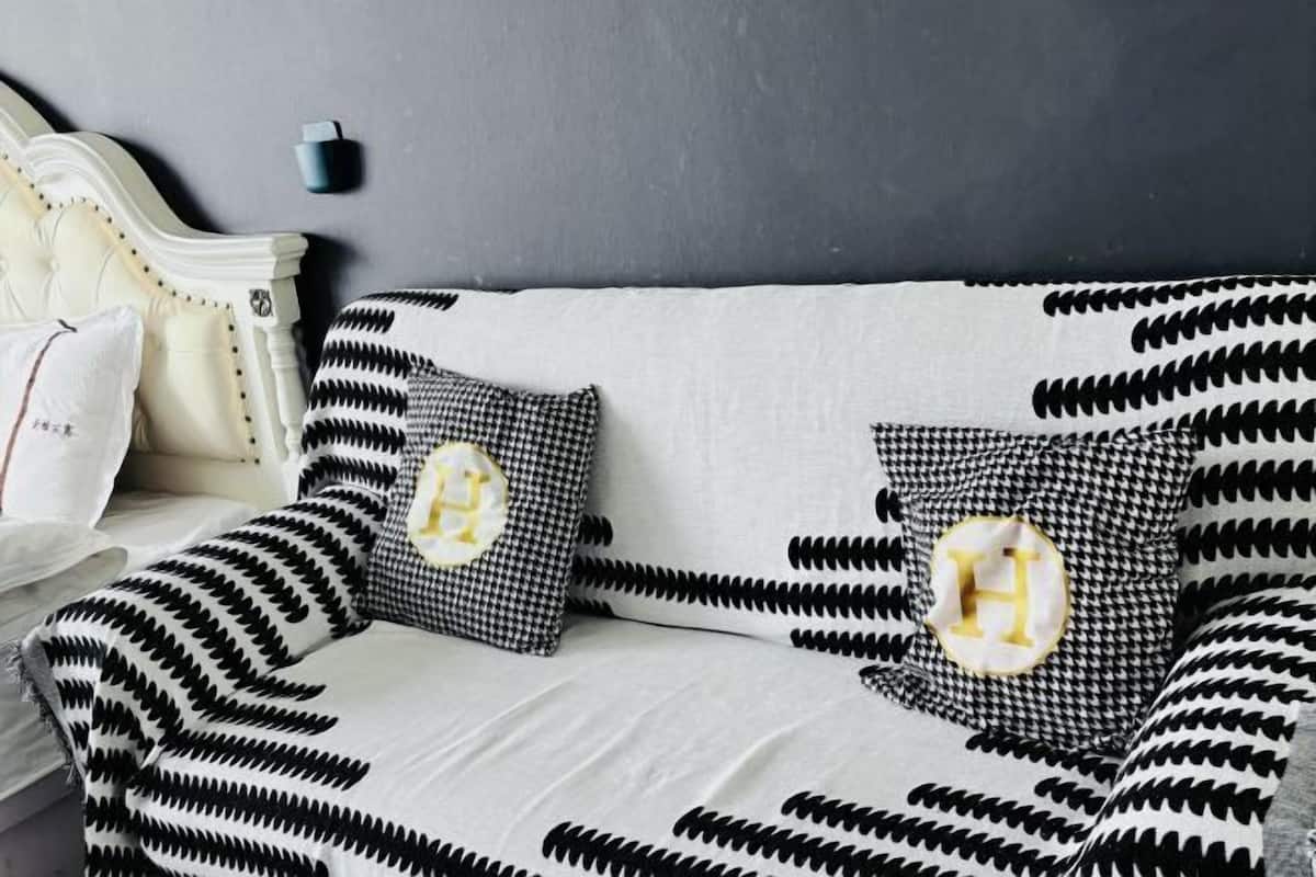Una cama con cabecera blanca, una colcha de diseño blanco y negro y dos almohadas con estampado pata de gallo en blanco y negro y una letra 'H' dorada.