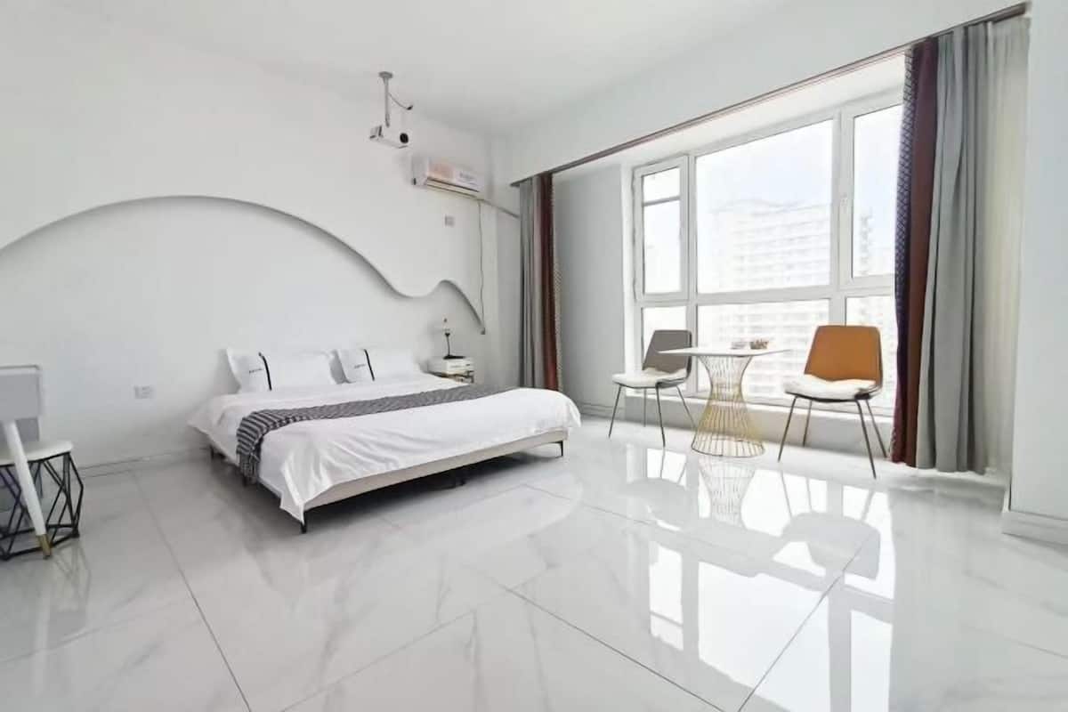 Una habitación de hotel moderna con una cama grande, una mesita pequeña, dos sillas, un escritorio y un ventanal con cortinas.