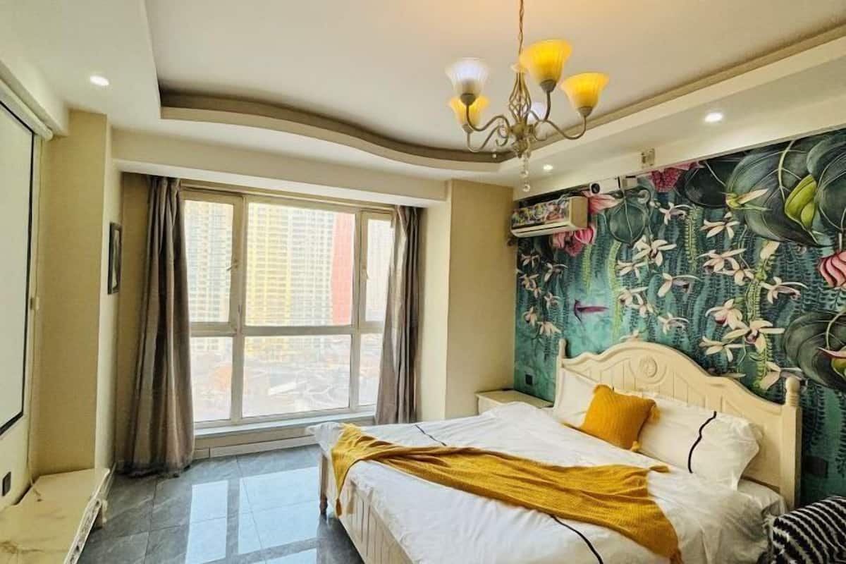 Una habitación de hotel con cama, ventana con cortinas, pared con un mural floral y un candelabro.