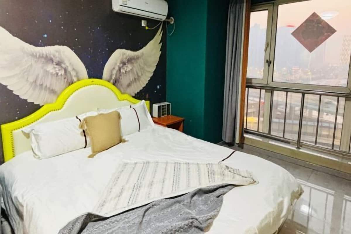 Una cama con cabecera blanca, una mesita de noche, un aire acondicionado montado en la pared y una ventana con vista a un paisaje urbano.