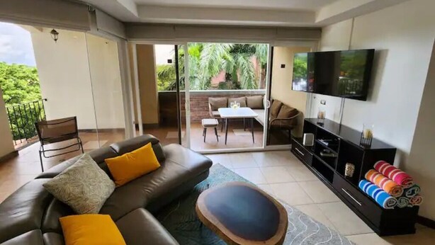 A living room with a sofa, coffee table, TV, and a balcony with a table and chairs. - Serenity Oasis in Playa Langosta (Playa Langosta)