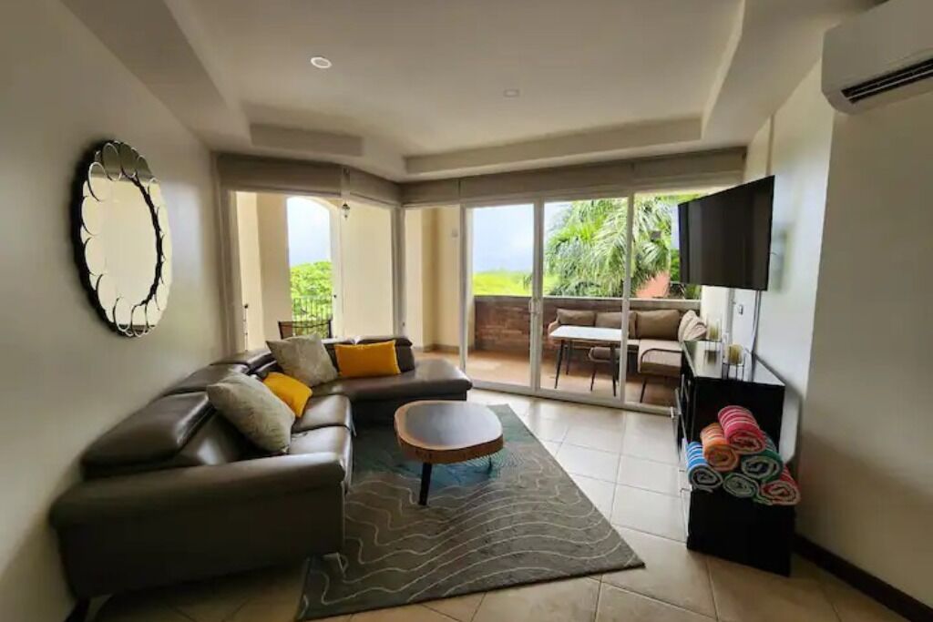 A modern living room with a sofa, a coffee table, and a TV. There is a balcony with a view of greenery.