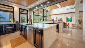A modern kitchen with dark cabinetry, a large island, and a dining area with a view of the outdoors. - Private Luxurious Beachfront Villa with Chef, Household Staff, Concierge  (Potrero)