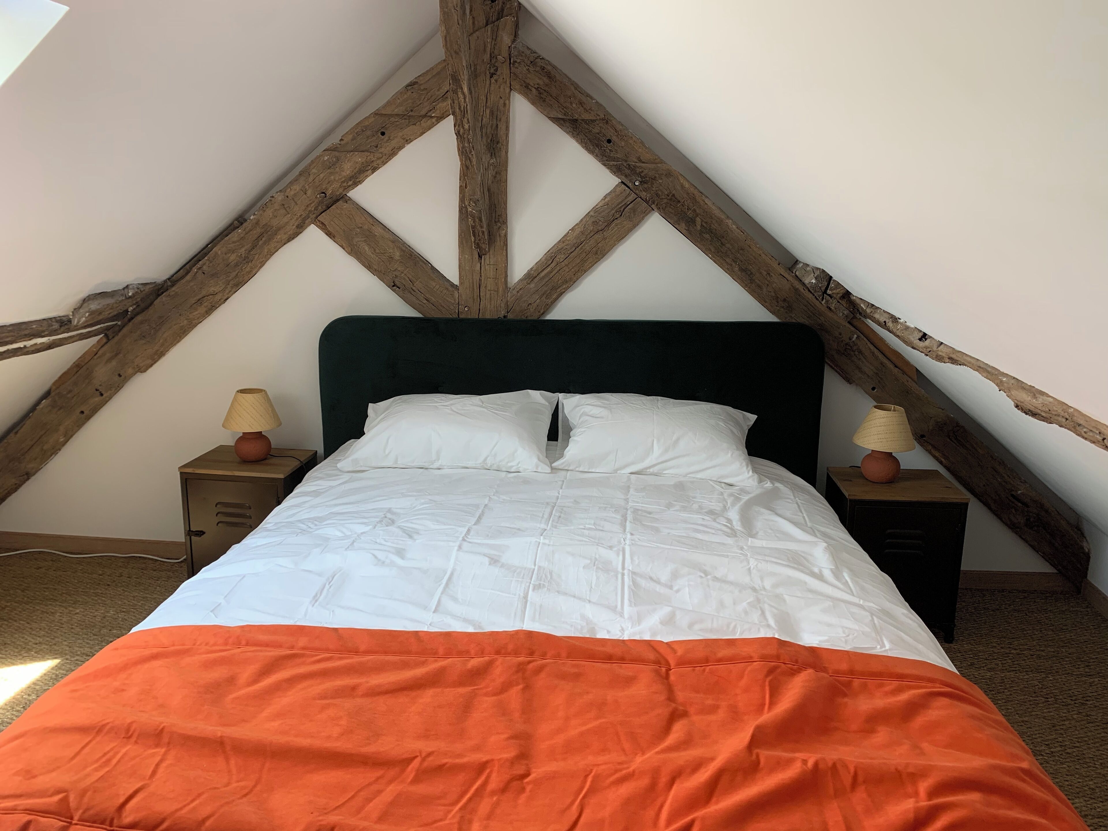 Un lit dans une chambre située sous les toits, avec un plafond en pente, des poutres en bois et deux tables de chevet équipées de lampes.
