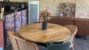 A round wooden table with a vase of flowers, surrounded by wicker and green upholstered chairs, set against a stone wall with a rustic kitchen in the background. - Gite Indépendant 4-6 Personnes Avec Piscine Privative (Gourdon)