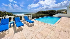 A pool area with a view of the ocean, a white fence, and blue lounge chairs. - Toes in the Sand | Beachfront Home & Private Pool (Christiansted)