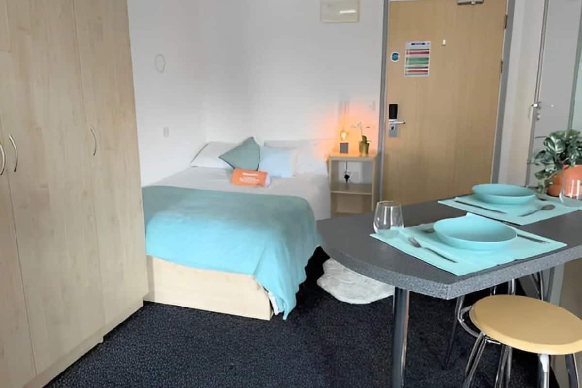 A small, neatly arranged room with a bed, a table with chairs, a nightstand, and a door.