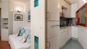 Una cocina compacta con muebles blancos, un refrigerador y un lavabo. En la habitación contigua hay una cama con almohadones estampados.