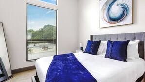 Een slaapkamer met een groot bed, een raam met zonwering, een kunstwerk aan de muur en een nachtkastje met een lamp.
