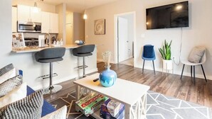 Een moderne woonkamer met een witte bank, een salontafel met gezelschapsspellen, een televisie en een keukenhoek met witte kasten en een magnetron.