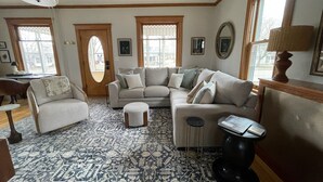 A living room with a sectional sofa, a round ottoman, and a coffee table. There are two windows with decorative glass panes, a door with a unique oval design, and a patterned rug on the floor. - Heron House - Elevated Vintage Downtown Victoria (Victoria)