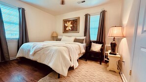 A bedroom with a bed, a nightstand, a lamp, a painting, and a window with curtains.