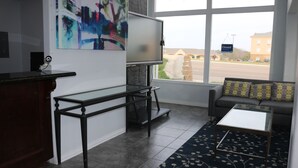 A modern living room with a glass coffee table, a sofa with patterned cushions, a TV on a stand, and a large window with a view of a parking lot and buildings. - MG INN & SUITES (Zapata)