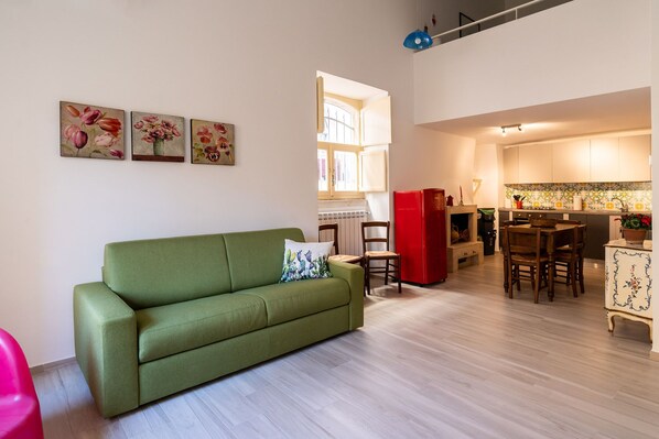 Ein Wohnzimmer mit einem grünen Sofa, einem Esstisch mit Stühlen, einem roten Kühlschrank und einer Küchenzeile mit Schränken und einer gefliesten Spritzschutzwand.