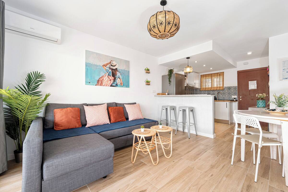 A modern living room with a grey sectional sofa, two round wooden stools, a white dining table with white chairs, and a kitchen area in the background.