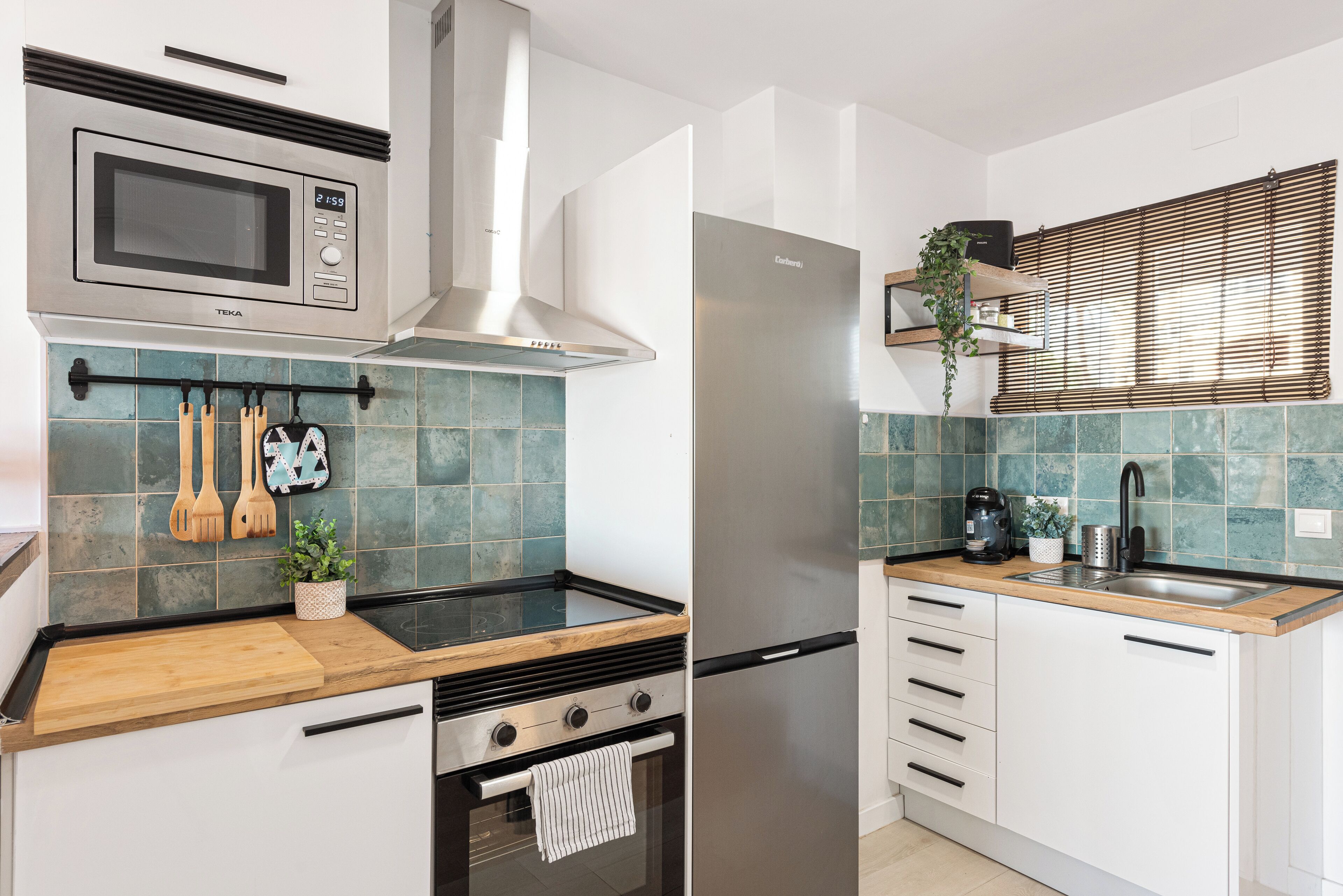 A modern kitchen with a microwave, stove, oven, and refrigerator. There are wooden utensils hanging on the wall, a plant, and a window with blinds.
