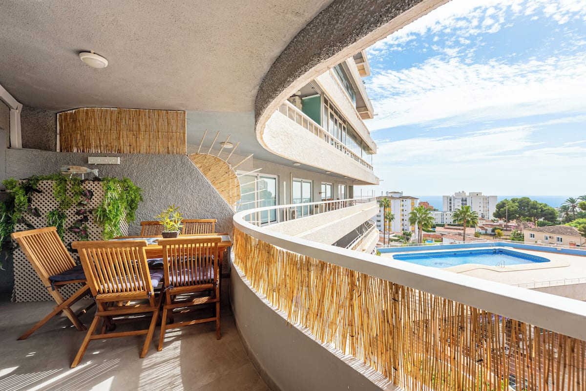 A balcony with a pool, wooden furniture, and a view of the cityscape.