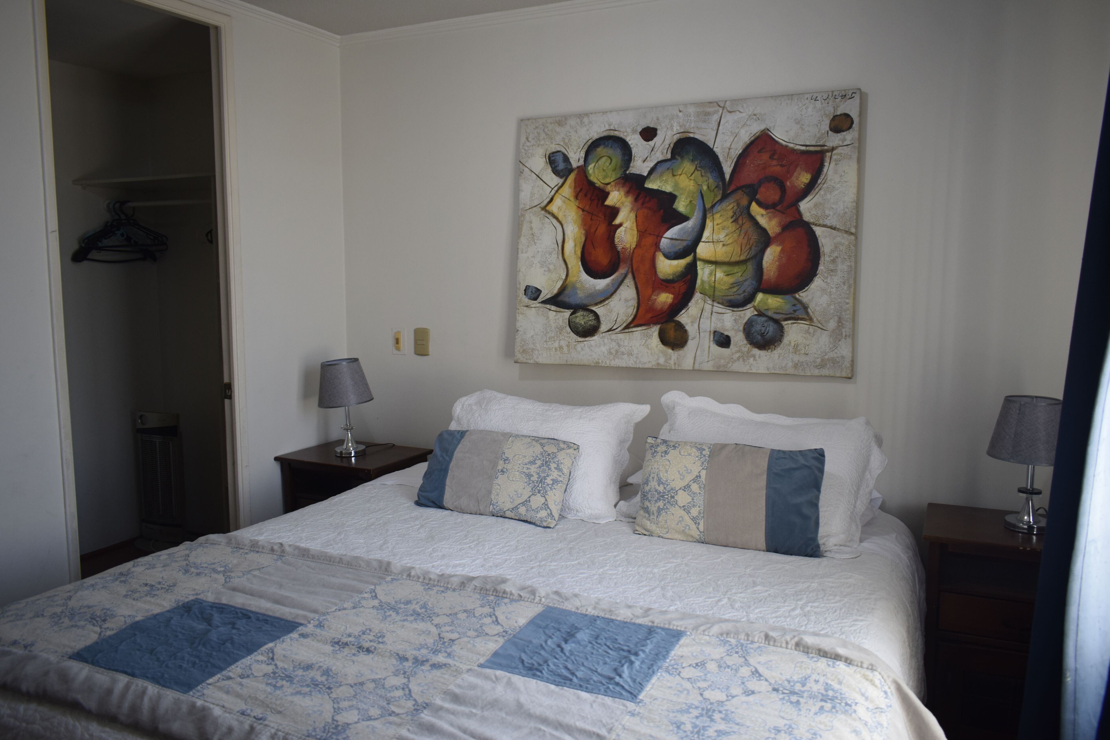 A neatly made bed with a quilt, a painting on the wall, and a closet with a hanger.