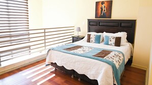 Quarto com cama, mesa de cabeceira, abajur, quadro e janela com persianas.