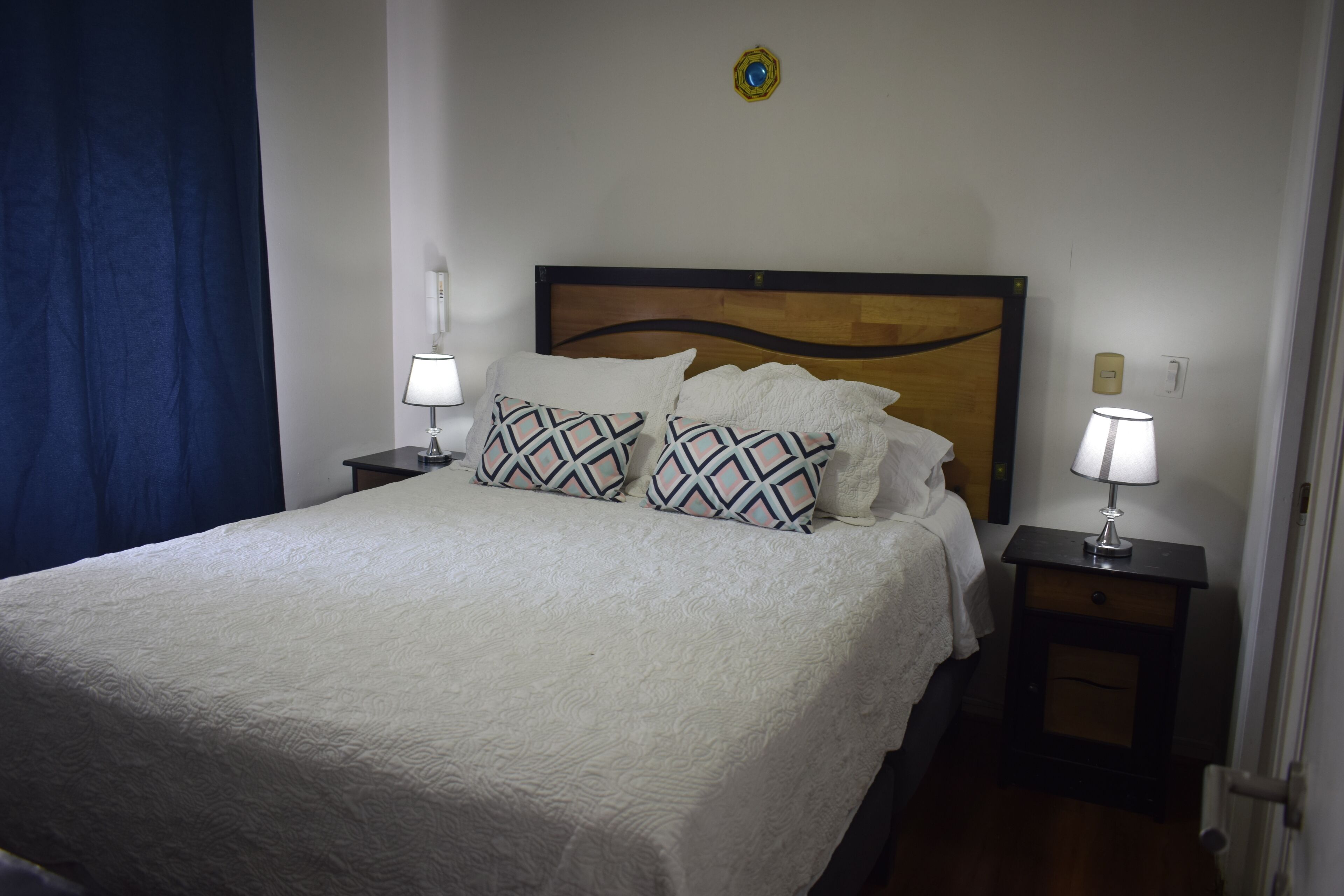 Un lit bien fait, recouvert d’une couvre-lit blanche et agrémenté d’oreillers à motifs, flanqué de deux tables de chevet avec des lampes, dans une pièce dotée d’une tête de lit en bois et d’un rideau bleu.
