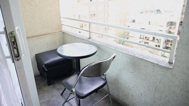 Uma varanda com uma mesa redonda pequena e duas cadeiras, uma porta e vista para prédios ao fundo.