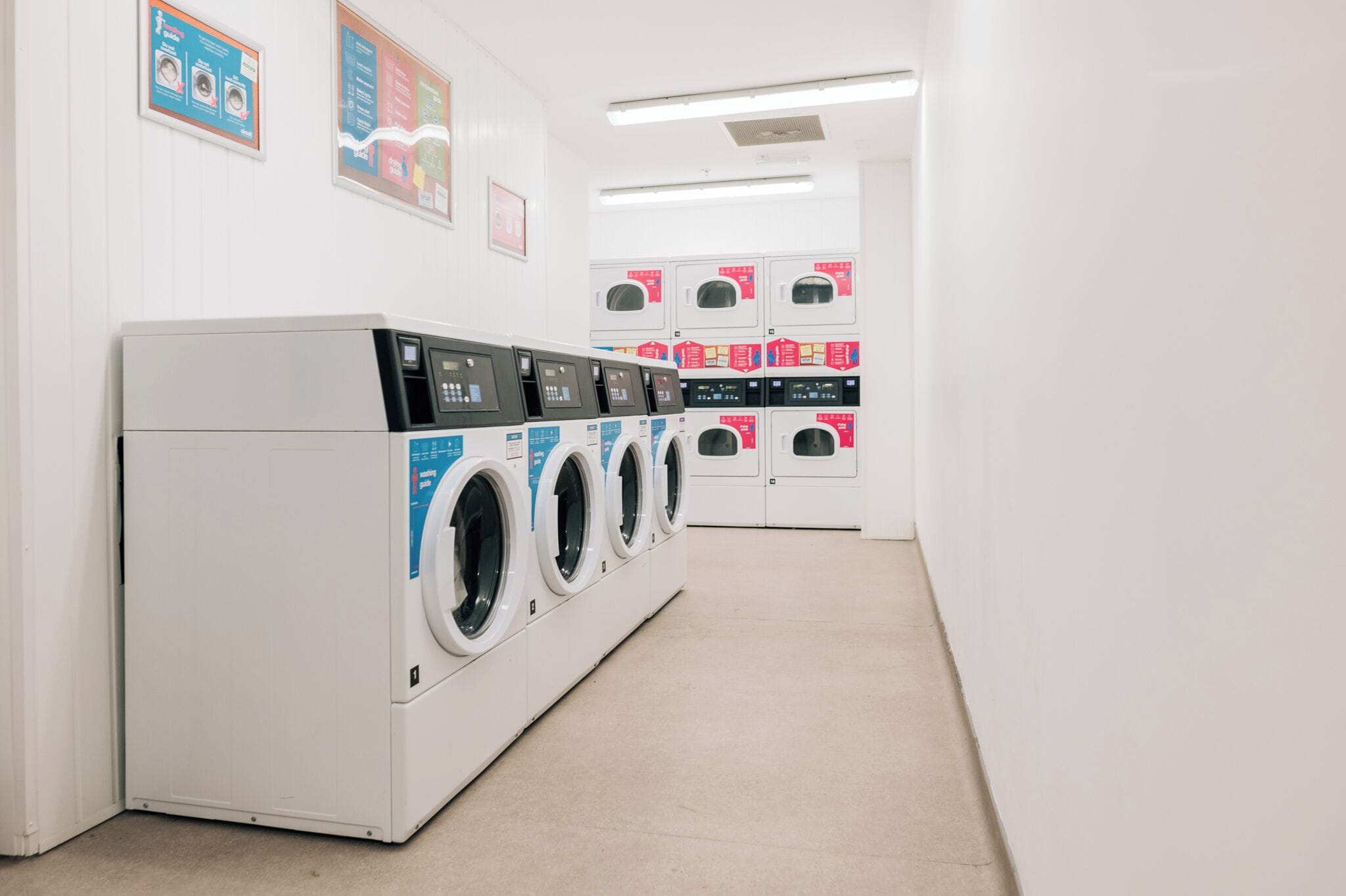 Une buanderie équipée de plusieurs machines à laver et de plusieurs séchoirs, avec un sol propre et des affiches informatives accrochées aux murs.