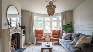 Ein Wohnzimmer mit Kamin, zwei karierte Sessel, ein graues Sofa, ein Holztisch und ein großes Fenster mit Vorhängen.