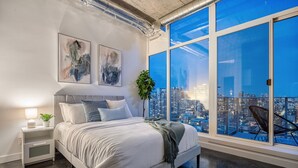 Ein modernes Schlafzimmer mit einem großen Bett, einem Nachttisch, einer Topfpflanze und Blick auf die Stadt durch große Fenster.
